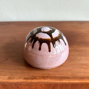 May include: A rounded, ceramic decorative object with a speckled, light pink base. The top features dark brown, dripped glaze details. The object is sitting on a wooden surface.
