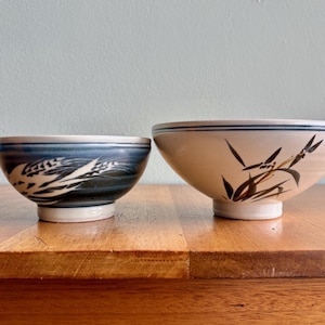 May include: Two ceramic bowls with decorative designs. The smaller bowl has a blue and white wheat pattern. The larger bowl is cream-colored with a bamboo design and blue trim. Both bowls are on a wooden surface.