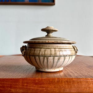 May include: A ceramic lidded jar with a round body and a lid with a small knob. The jar is beige with vertical green stripes and small handles. The jar is sitting on a wooden surface.
