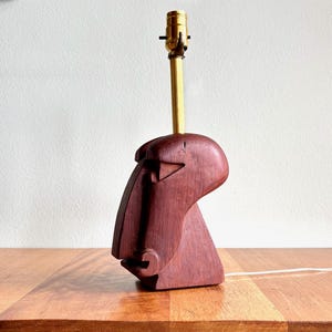 May include: A wooden table lamp with a carved face design. The lamp has a reddish-brown finish and a brass-colored neck and lamp holder. The lamp is plugged into an electrical outlet.
