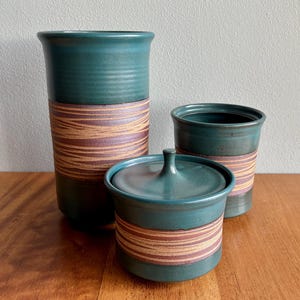 May include: A set of three teal ceramic containers with a brown and purple striped design. The set includes a tall vase, a small lidded jar, and a shorter cup. The containers are arranged on a wooden surface.