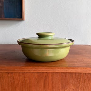 May include: A round, light green ceramic casserole dish with a matching lid. The dish features a smooth, glossy finish and a dark brown rim along the edge of the lid and the top of the bowl. It has a small, circular knob handle on the center lid.