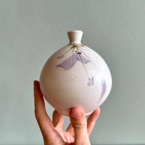 May include: A white ceramic vase with a bulbous body and a narrow neck. The vase is decorated with a floral design in shades of purple and green. The vase is approximately 5 inches tall.
