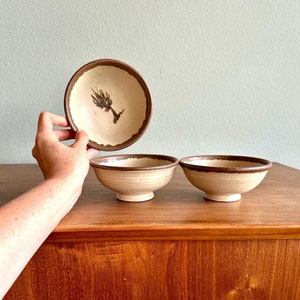 May include: Three ceramic bowls with a beige interior and a dark brown rim. One bowl is held up, revealing a brown floral design. The bowls sit on a wooden surface.
