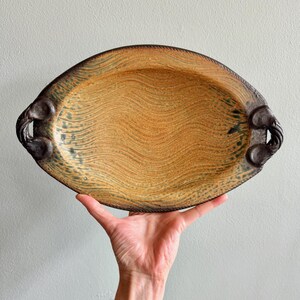 May include: An oval ceramic serving platter featuring a textured, wavy pattern in shades of golden tan and brown. The platter has two dark, sculpted handles on opposite sides with a rope-like border detail along the outer edge of the rim.