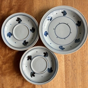 May include: Three ceramic plates with a light blue background and dark blue floral designs. The plates vary in size and feature a central circle and dotted border. The plates are arranged on a wooden surface.
