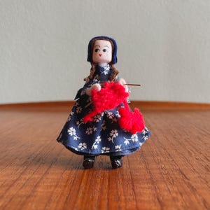 May include: A miniature doll dressed in a navy blue dress with white floral patterns and a blue bonnet. The doll is holding a red yarn ball and knitting needles, depicting a crafting theme. The doll is standing on a wooden surface.
