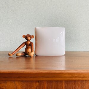 May include: A wooden monkey figurine with articulated limbs sits next to a white, square vase. The monkey is brown with light tan accents on its hands and feet. The vase has a smooth, glossy finish.