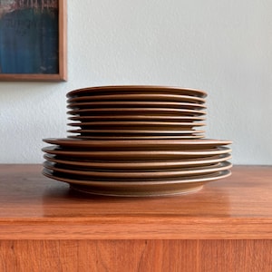 May include: A stack of brown ceramic plates on a wooden surface. The plates are arranged in two stacks, one larger than the other. The plates have a simple, minimalist design and a matte finish. The background includes a painting and a white wall.