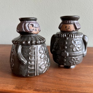 May include: Two ceramic figurines, each with a dark gray glaze. The figures have stylized faces and are dressed in decorative outfits with floral patterns. One has a ruffled collar and the other has a more structured design. They are displayed on a wooden surface.