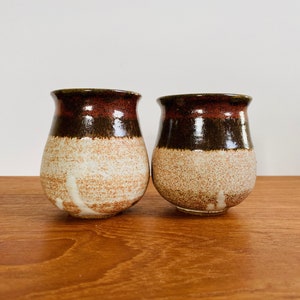 May include: Two handmade ceramic tumblers. Each tumbler has a dark brown rim, a band of reddish-brown, and a speckled tan body. The tumblers are displayed on a wooden surface against a white background. The pottery has a rustic, handcrafted aesthetic.
