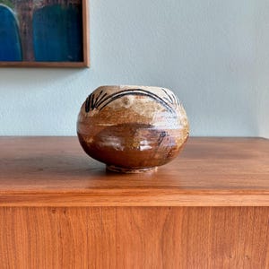 May include: A handcrafted, round ceramic bowl with a brown and tan mottled glaze. The bowl features a dark, linear design around the rim. The bowl is displayed on a wooden surface, showcasing its rustic aesthetic.