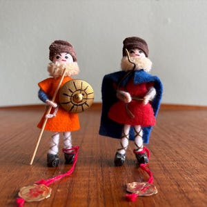 May include: Two handmade miniature warrior dolls. One doll wears an orange tunic, blue sleeves, and a brown hat, holding a spear and a gold shield. The other doll wears a red tunic and a blue cape, holding a bow. Both dolls have white fur trim and black shoes.