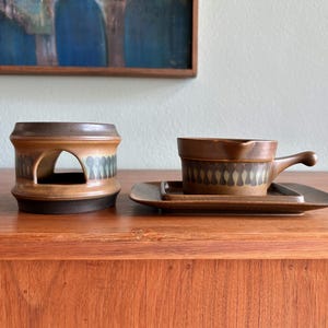 May include: A brown ceramic soup bowl with a handle, resting on a matching rectangular plate. Beside it is a brown ceramic warmer with arched openings and a decorative pattern. The items are on a wooden surface.