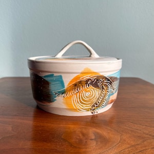May include: A ceramic lidded jar with a cream-colored base. The jar is decorated with abstract brushstrokes in blue, brown, and orange. A bird design is featured on the front. The lid has a small handle.