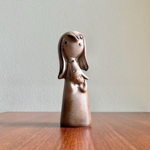 May include: A ceramic figurine of a girl holding a bird. The figurine is a light brown color with black details. The girl has long hair and is wearing a dress. The figurine is standing on a wooden surface.