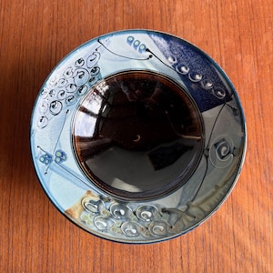 May include: A handmade ceramic plate with a dark brown center and a light blue rim. The rim features decorative patterns in white and blue, including swirls and dots. The plate is set against a wooden surface.