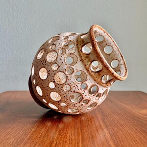 May include: A ceramic vase with a speckled, off-white and brown finish. The vase has a rounded body with numerous circular cutouts and a flared rim. The base is a dark brown color. The vase is displayed on a wooden surface.
