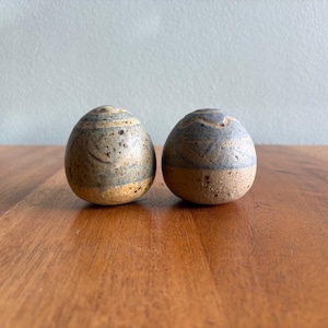 May include: Two handcrafted ceramic egg-shaped sculptures. Each has a speckled, textured surface with bands of gray and tan. The eggs are displayed on a wooden surface, showcasing their unique, organic forms. These decorative pieces add a touch of rustic charm.