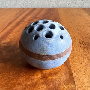 May include: A blue ceramic flower frog with multiple holes for arranging stems. The spherical frog has a brown band around the middle. The flower arranging tool is on a wooden surface.