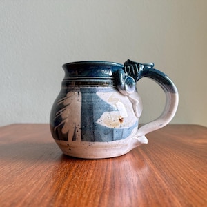 May include: Handmade ceramic mug with a unique design. The mug features a blue rim and handle, with a white and blue pattern on the body. The handle has an artistic, sculpted detail. A functional and decorative piece.