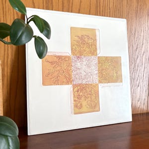 May include: A square white art print featuring a central cross-shaped design in shades of yellow and orange. The cross is filled with intricate, fine-line botanical illustrations of flowers and foliage, creating a detailed, layered visual texture.
