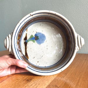 May include: A round ceramic bowl with a speckled white interior and a blue floral design. The rim features blue and brown stripes, with ribbed handles on either side. The bowl is set on a wooden surface.