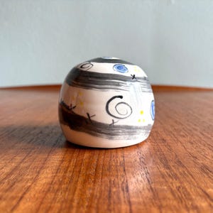 May include: A small, rounded ceramic container with a cream-colored base. It features black horizontal stripes, blue and yellow dots, and black spiral and cross designs. The container sits on a wooden surface.
