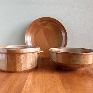 May include: A set of three ceramic serving dishes in shades of brown. The set includes a round plate, a bowl, and a casserole dish with handles. The dishes have a rustic, earthy appearance, suitable for serving food.