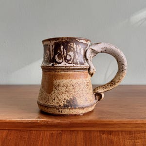 May include: A ceramic mug with a speckled beige and brown glaze. The upper section features a dark brown band with decorative circular patterns, while the lower body has a textured, mottled finish. It includes a curved, speckled handle.