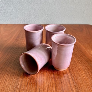 May include: A set of four cylindrical ceramic cups with a smooth, glossy finish in varying shades of soft pink and mauve. The cups feature a simple, handleless design with a slightly flared rim and a visible unglazed base on the bottom.