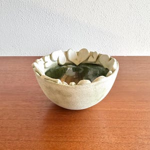 May include: A handmade ceramic bowl with a heart-shaped rim. The bowl has a light beige exterior and a dark green interior. The bowl is sitting on a wooden surface.