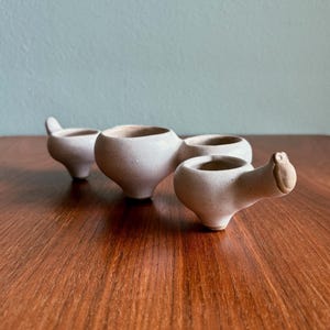 May include: Four small, off-white ceramic pots with a speckled texture. One pot has a handle, and another has a small, animal-like head. The pots are arranged on a wooden surface, with a light blue background.