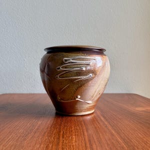 May include: A round ceramic jar with a lid, featuring a glossy glaze in shades of brown, tan, and cream. The exterior surface displays a swirling, abstract pattern of raised white lines that wrap around the body of the vessel.