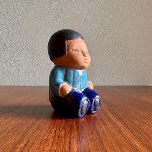 May include: A ceramic figurine of a seated figure with black hair, wearing a blue shirt and dark blue pants. The figurine has a light brown face and is sitting on a wooden surface.