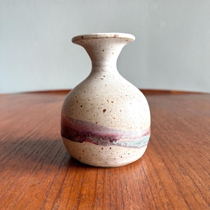 May include: A small, speckled ceramic vase with a bulbous body and flared neck. The vase features a band of burgundy and gray around the middle. The neutral tones and unique shape make it a decorative piece.