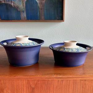 May include: Two deep blue ceramic bowls with integrated side handles and matching lids. The lids feature a cream-colored base with a detailed floral pattern in shades of blue, green, and yellow. The bowls have a smooth, matte finish.