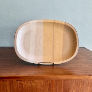 May include: An oval ceramic platter featuring a speckled finish with vertical color-blocked stripes in shades of cream, light beige, and golden yellow. The dish has a smooth, rounded rim and is displayed upright on a small metal stand.