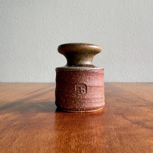 May include: A handcrafted, brown ceramic vase with a textured surface. The vase has a cylindrical body with a slightly flared top and a dark brown, rounded neck. A square symbol is imprinted on the front. The vase sits on a wooden surface.