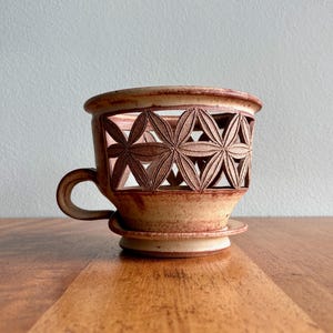 May include: A handmade ceramic coffee dripper with a unique floral cut-out design. The dripper is a warm beige color with brown accents and a small handle. It sits on a matching saucer, perfect for brewing coffee.