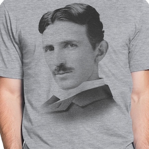 May include: A black and white photograph of Nikola Tesla, a famous inventor and engineer, wearing a suit and tie. The image is printed on a heather grey t-shirt.