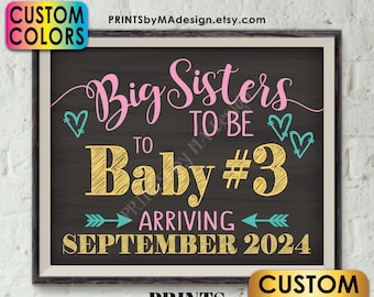 May include: A chalkboard style sign with the text "Big Sisters to be Baby #3 Arriving September 2024". The sign is decorated with pink and teal hearts and arrows. The text is in a yellow, pink, and teal font.