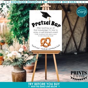 May include: A graduation-themed sign on an easel. The sign reads "Pretzel Bar" with text below, a graduation cap graphic, and a pretzel illustration. The sign is on a wooden easel, with floral arrangements in the background.
