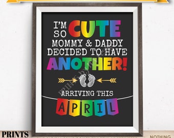 Könnte beinhalten: Ein rechteckiger Digitaldruck in einem dunklen Rahmen mit dem Text I'M SO CUTE MOMMY & DADDY DECIDED TO HAVE ANOTHER! in Regenbogenfarben und Weiß. Darunter ein goldener Pfeil, weiße Babyfußabdrücke und der Text ARRIVING THIS APRIL.