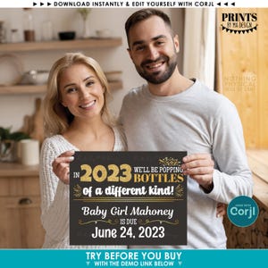 May include: A couple smiles, holding a black sign with gold text announcing a baby girl is due June 24, 2023. The sign reads "We'll be popping bottles of a different kind in 2023." The design is from Prints by MA Design.
