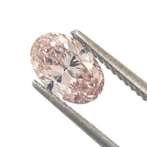 Könnte beinhalten: Ein oval geschliffener loser Edelstein mit blassrosa Farbton und einer facettierten Oberfläche, die das Licht reflektiert. Der Stein wird von einer silberfarbenen Metallpinzette gehalten und zeigt seine Transparenz und Symmetrie vor weißem Grund.
