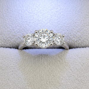 May include: A three-stone ring featuring a large round-cut center diamond flanked by two smaller round-cut diamonds. The stones are set in a polished silver-toned metal band with four-prong settings for each stone, creating a classic, symmetrical look.