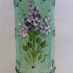 Puede incluir: Una botella de vidrio verde con un dise&ntilde;o pintado a mano de flores moradas y blancas. La botella tiene una superficie texturizada y una tapa de metal.