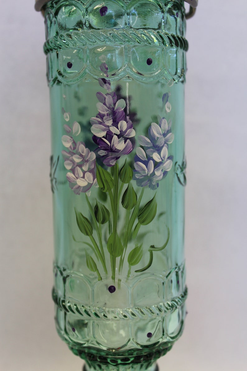 Puede incluir: Una botella de vidrio verde con un dise&ntilde;o pintado a mano de flores moradas y blancas. La botella tiene una superficie texturizada y una tapa de metal.
