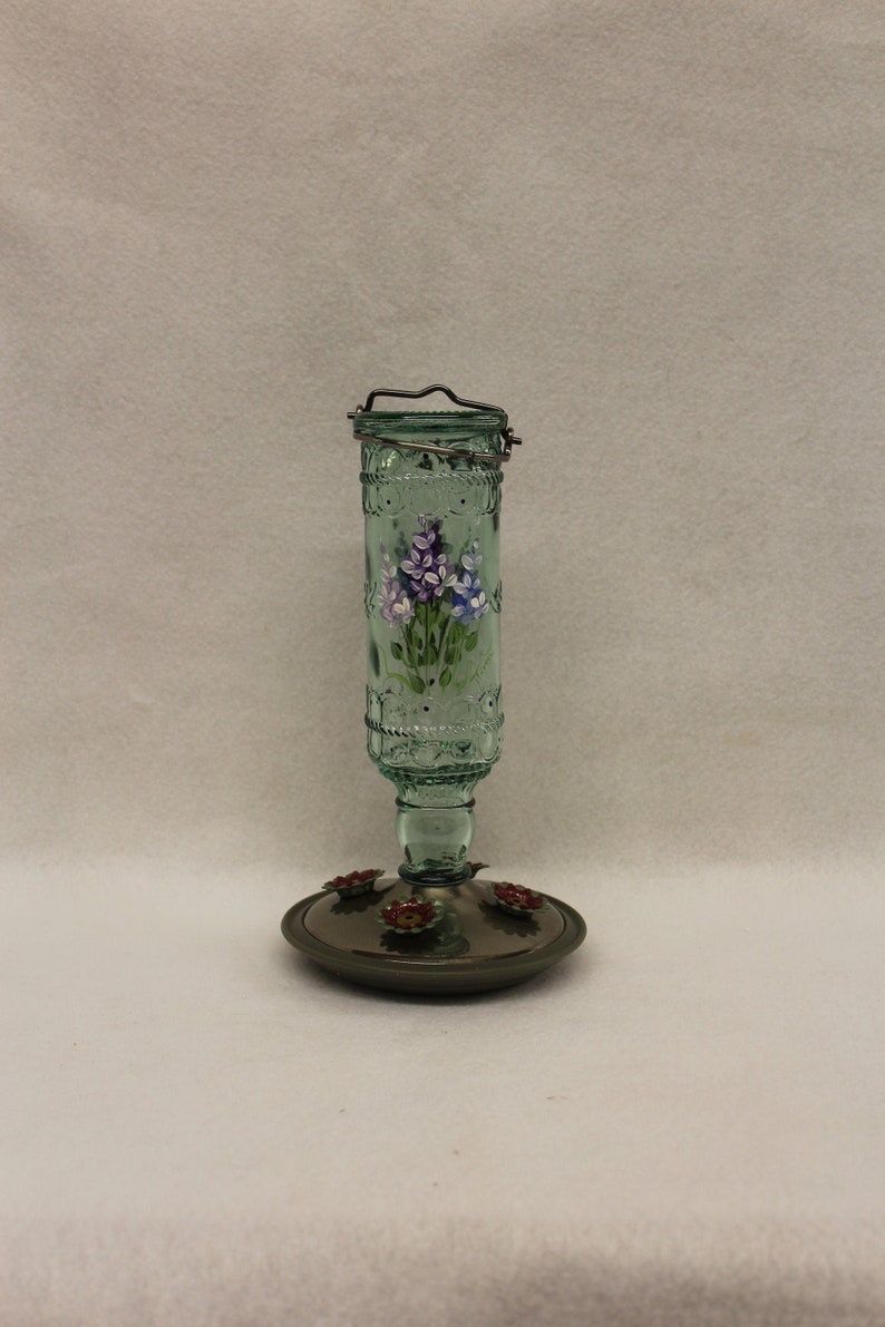 Puede incluir: Un comedero para colibr&iacute;es de vidrio verde con un colgador de metal y un dise&ntilde;o floral pintado. El comedero tiene una base de metal con acentos de flores rojas.